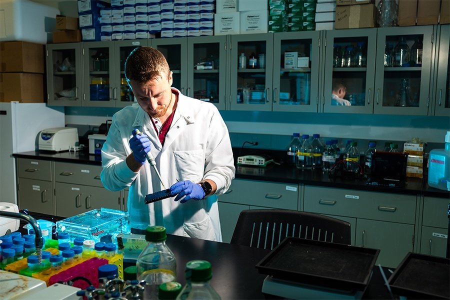 Male in a white lab coat in scientific lab uses a pipette on samples.