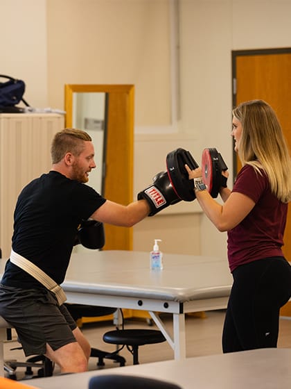 A pre-physical therapy student works with a patient to build strength through boxing work with pads.