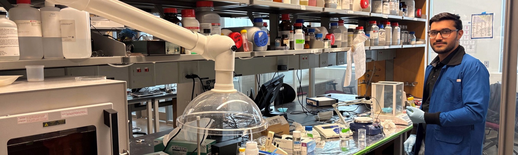 an engineering student in a lab coat stands next to counter filled with instruments in a lab setting
