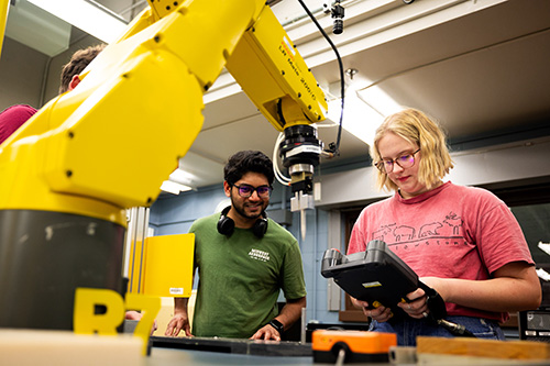 Two students using a hand held computer interface, programming a yellow robotic arm.