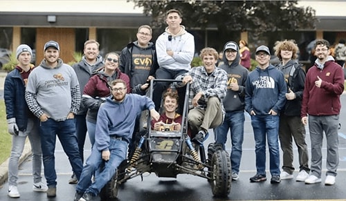 A group of students standing around one of the Baja cars in a parking lot.