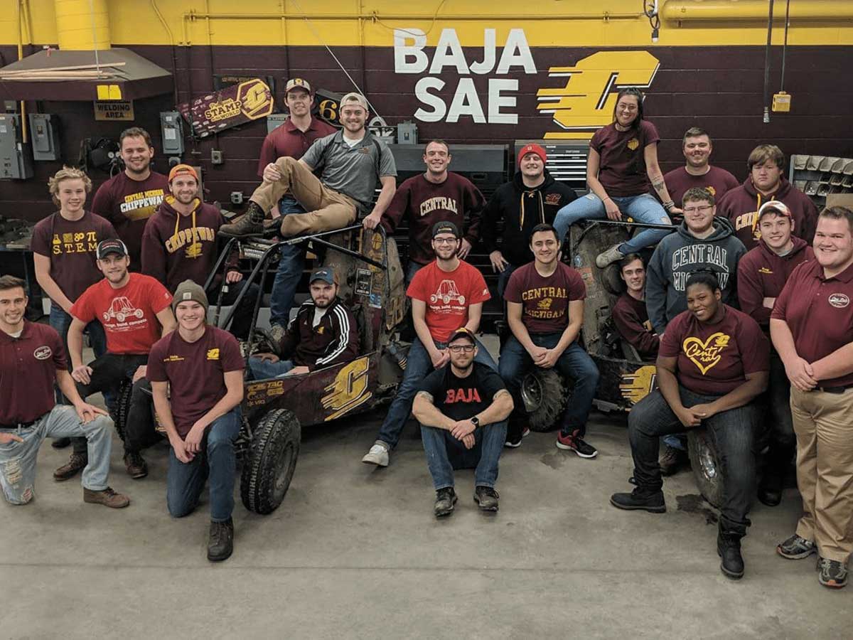 A group people wearing maroon shirts stand in a garage around two baja cars.