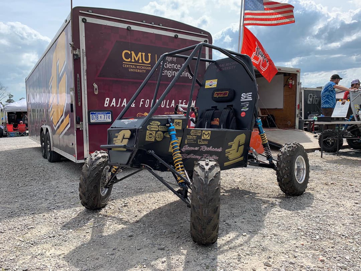 The CMU Baja Race Car in front of the CMU branded trailer with flags flying in the background.