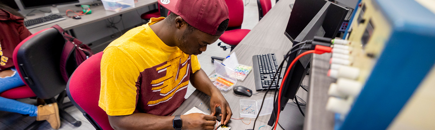 Electrical Engineering student in a CMU shirt and hat working on a circuit board in front of electrical equipment.