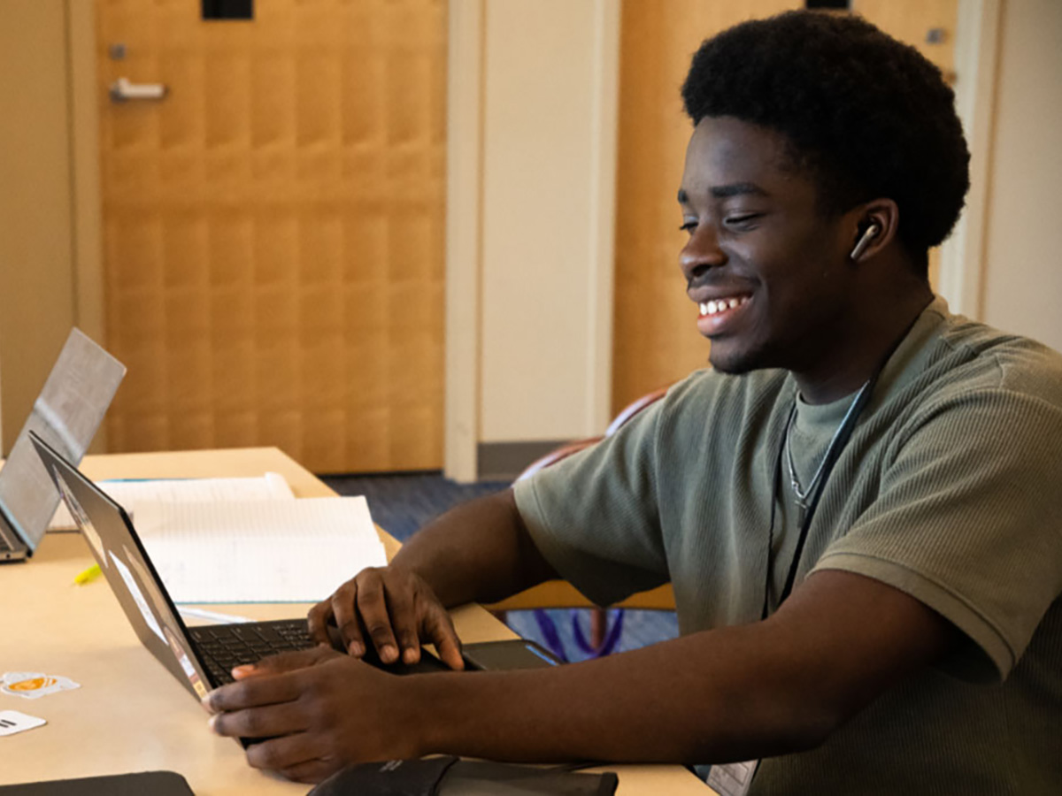 A young man smiles while looking at the screen of his laptop.