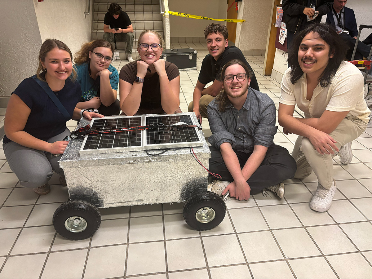 A group of students posing around a silver box with solar panels on top.