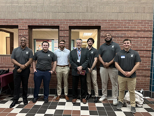 Seven students and faculty stand together in a hallway for a photo