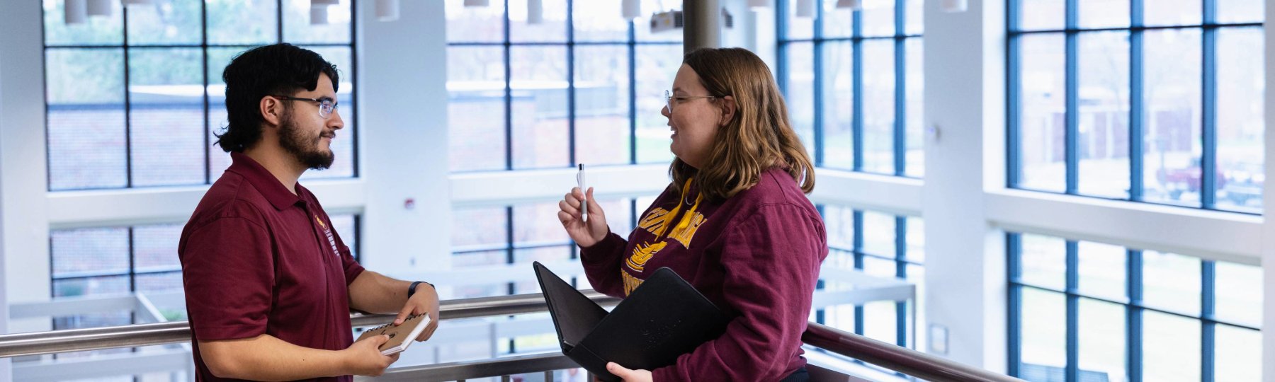 Two students talk and smile while standing in an academic building with large windows. One holds a notebook, and the other holds a laptop and pen. Both wear CMU-branded maroon clothing, suggesting a school-related discussion.
