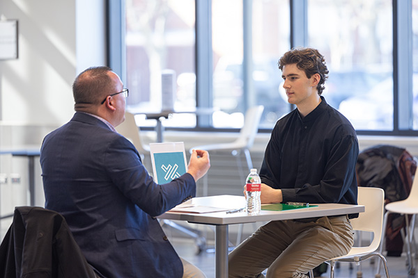 A student sitting at a table speaking with a man in a suit.
