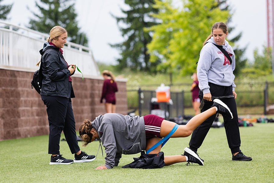 Two athletic training students stand watching an athlete do leg exercises on the field.