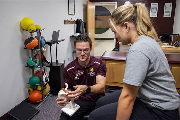 Athletic Training Student in the ICC 700x467 An athletic training student sitting in the Injury Care Center holds an anatomical model and talks with a patient.