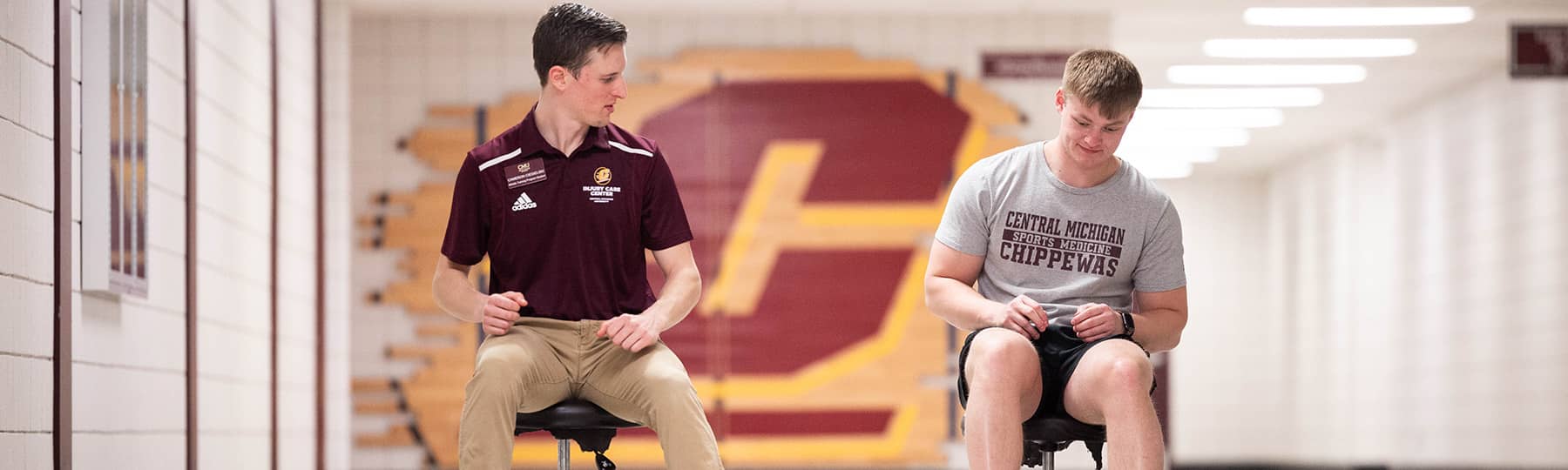 Injury Care Center Rehabilitation 1800x540 An athletic training student wearing a name tag, maroon polo, and khaki pants sits on a stool looking at a patient in a gray t-shirt sitting on a stool in the hallway outside of the Injury Care Center.