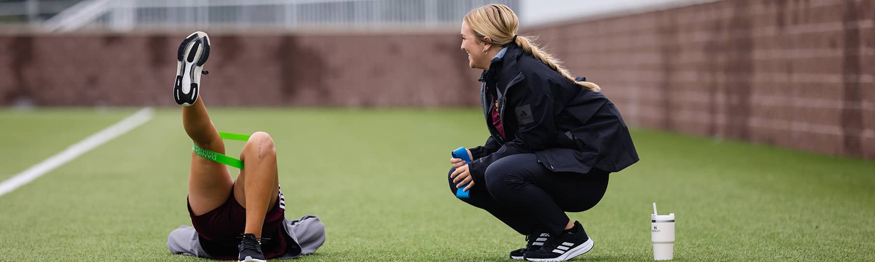 An athletic training student wearing a black jacket and black pants and holding a blue band squats near a soccer athlete who is laying on a green turfed surface holding a one-legged bridge position with a green band around their thighs.