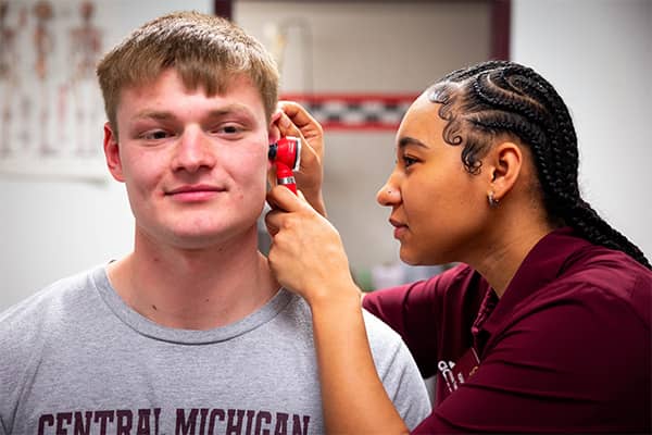 An athletic training student wearing a maroon polo stands near a patient wearing a gray t-shirt. The athletic training student holds the patient's ear and looks inside the ear with an otoscope.