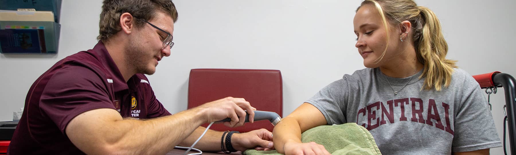 An athletic training student wearing a maroon polo and glasses sits in the Injury Care Center holding an ultrasound wand on a patient's right forearm that is resting on an olive colored towel. The patient is wearing a grey t-shirt.
