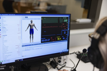Computer monitor displaying healthcare simulation software with a virtual patient model and real-time vital sign data, including heart rate, waveform monitoring, and oxygen saturation.
