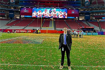 Sport management student, Quintin Hunsinger, stands on the State Farm Stadium football field covered in red and yellow confetti with the Super Bowl 57 Champions, Kansas City Chiefs football players pictured on the jumbotron behind him in the endzone.