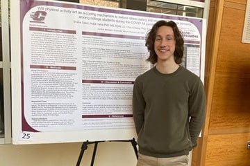 Athletic training and nutrition student, Shane Salski, stands with his hands behind his back, smiling at the camera in front of his research poster in the atrium of the College of Health Professions.