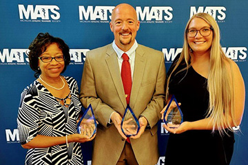 Rene in a black and white dress, Blain in a tan suit jacket and maroon tie, and Kayla in a black dress hold their awards while smiling at the camera.