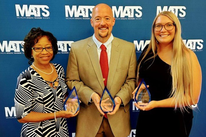 Rene in a black and white dress, Blain in a tan suit jacket and maroon tie, and Kayla in a black dress hold their awards while smiling at the camera.