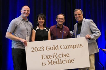 Dr. Saltarelli in a maroon polo poses between three Exercise is Medicine representatives dressed in gray and black holding an Exercise is Science 2023 Gold Campus Award banner.