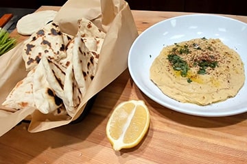 A basket of homemade pita bread, a plate of hummus, and a fresh cut lemon on a cutting board..