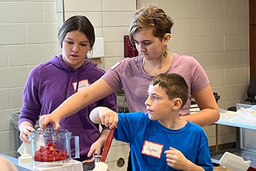 Three children use a blender as they participate in a cooking demonstration event on campus.