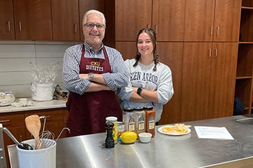 Male professor and a female student pose with arms-folded behind a cooking station in the campus test kitchen.