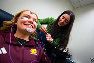 A student standing behind another student with electrodes on their face.