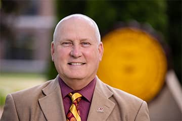 Greg Zimmerman, wearing a maroon collared shirt, CMU tie, and brown suit coat, stands outside in front of the CMU seal in Warriner Mall and smiles at the camera.