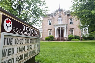 The Ionia Community Library sign with three listed events sits in front of two green trees and a tan two-story brick building.