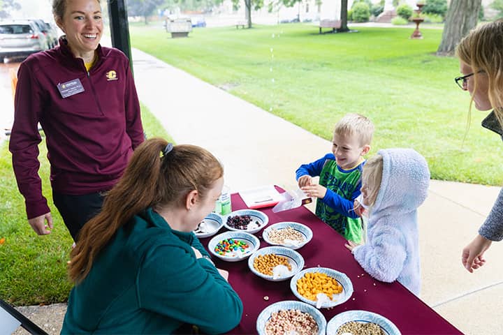A boy and a girl in a purple hood stand in front of a maroon table with snacks in eight paper bowls. Both are smiling at a student in a turquoise jacket sitting behind the table. Rachael Nelson is smiling in a maroon jacket while standing near the table.