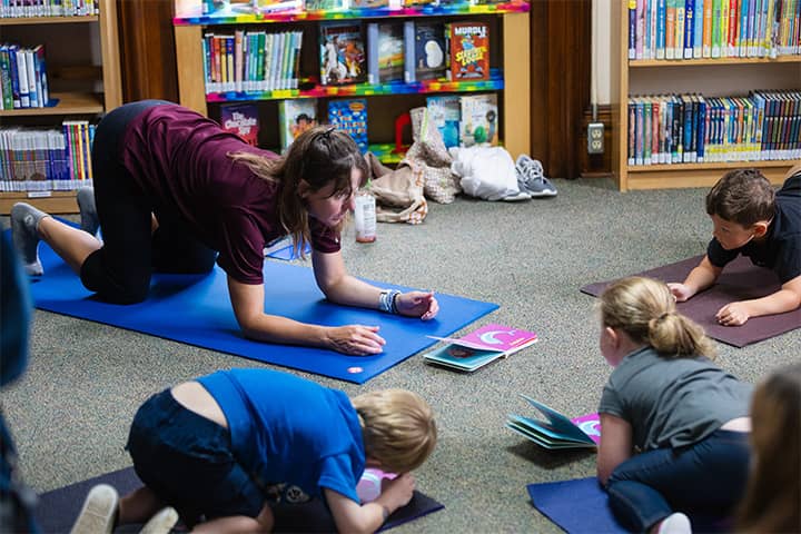 Jennifer Jones in a maroon shirt teaches a yoga pose on a blue mat to three young children on their own yoga mats in front of her. Jennifer is looking at the children while they are looking at her. Between them lay two open Yoga Whale books.