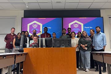 A group of sixteen students and faculty pose together smiling while standing between a wooden podium supporting computer monitors and two videos screens displaying purple, blue, and yellow graphics.