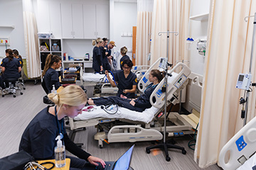 Ten nursing students dressed in dark gray scrubs practice taking vital signs in the simulation lab. One student sits at the front of the lab working on a laptop while others are standing or sitting in the background.