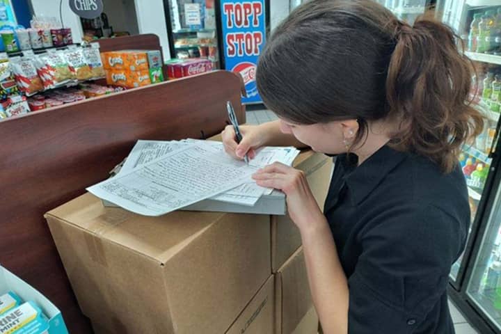 Linday Uzarski, wearing a black polo, stands near a stack of cardboard boxes where she rests her clipboard and papers. She is filling out paperwork in a convenience store where she is surrounded by snacks and bottled drinks.