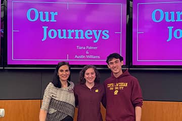 Two students dressed in maroon and a professor wearing a gray sweater stand together in front of a maroon screen with the title Our Journeys on it.