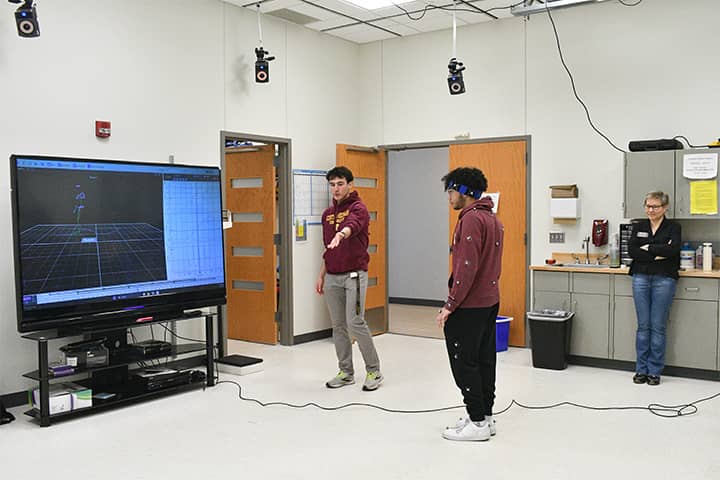 A student in a maroon sweatshirt directs another student who is standing front of a television screen with sensors placed on his sweatshirt and pants. Dr. Ustinova stands watching the students with her arms folded.