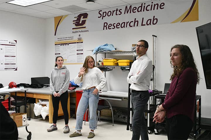 Two students stand to the right and one to the left of Dr. Zuhl in the Sports Medicine Lab in the College of Health Professions. Research posters and banners are fixed to the wall and supplies are positioned behind them.