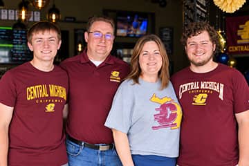 Kip and Nicole Ferguson stand together smiling with their sons Thomas and Ryan at their sides. Thomas, Ryan, and Kip are wearing maroon Central Michigan University shirt while Nicole is wearing a gray Central Michigan University shirt.
