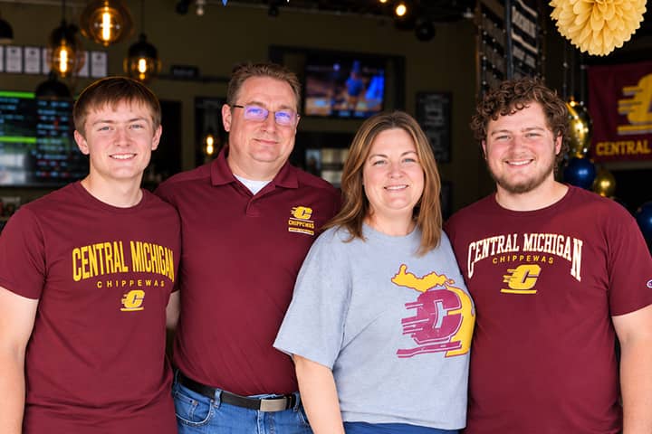 Ferguson Family 720x480 Kip and Nicole Ferguson stand together smiling with their sons Thomas and Ryan at their sides. Thomas, Ryan, and Kip are wearing maroon Central Michigan University shirt while Nicole is wearing a gray Central Michigan University shirt.