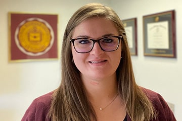 Beth wears a maroon sweater and glasses while looking at the camera in her office.