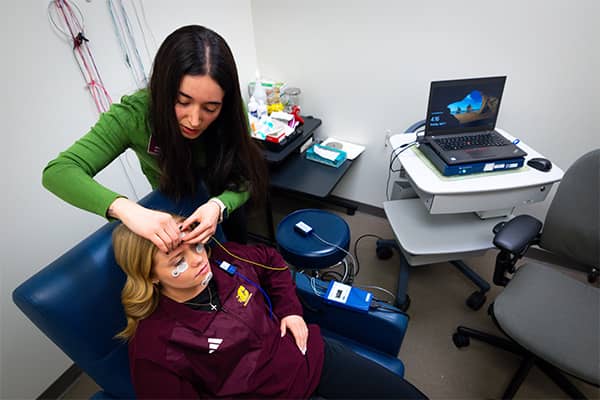 An audiology student wearing a green sweater stands and places a small patch on a patient's forehead. The patient is sitting in a blue chair wearing a maroon CMU quarter zip sweatshirt.