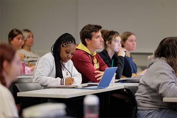 Four students sit together at a table in an American Sign Language class. Three students look forward while the fourth student holds a pencil and writes in a notebook set behinds an open blue laptop.