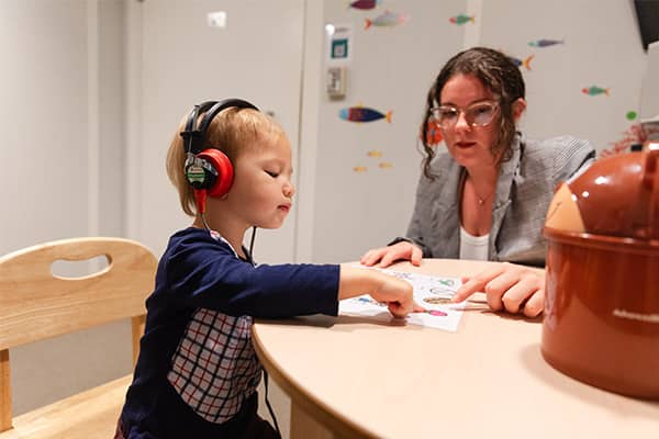 An audiology student wearing glasses and a gray blazer sits at a children's table next to a toddler wearing headphones. Both are pointing at pictures on a piece of paper on the table.