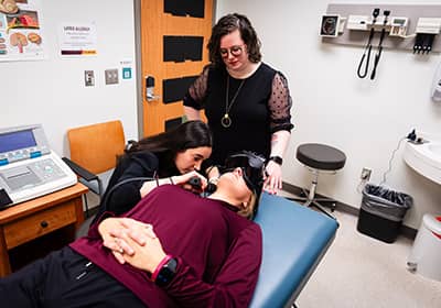A patient in a maroon top lies on an exam table wearing large goggles. An audiology student dressed in black sits next to the patient and looks inside their ear using an otoscope. An audiologist dressed in black stands to oversee the audiology student.
