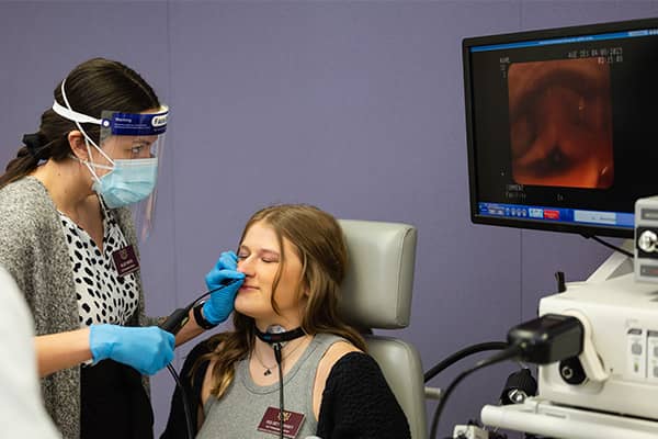 A patient is sitting in a chair and a speech-language pathology student wearing gloves, a face mask, and face shield stands near looking at a computer screen while guiding a videostobe into the patient's nose.