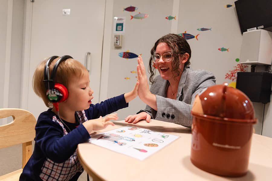 A Speech Language Pathology student where glasses and a gray blazer and a toddler where black and red headphones high five each other.