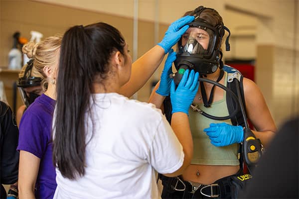 A student with her hair in a pony tail wearing a white t-shirt and blue gloves holds a full-face respirator in place on her partner who is wearing blue gloves and an olive green tank top.