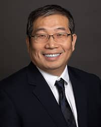Way Ge 200x250 Way Ge, wearing a dark blue tie, white collared shirt, and black suit coat, stands in front of a dark gray backdrop and smiles at the camera.